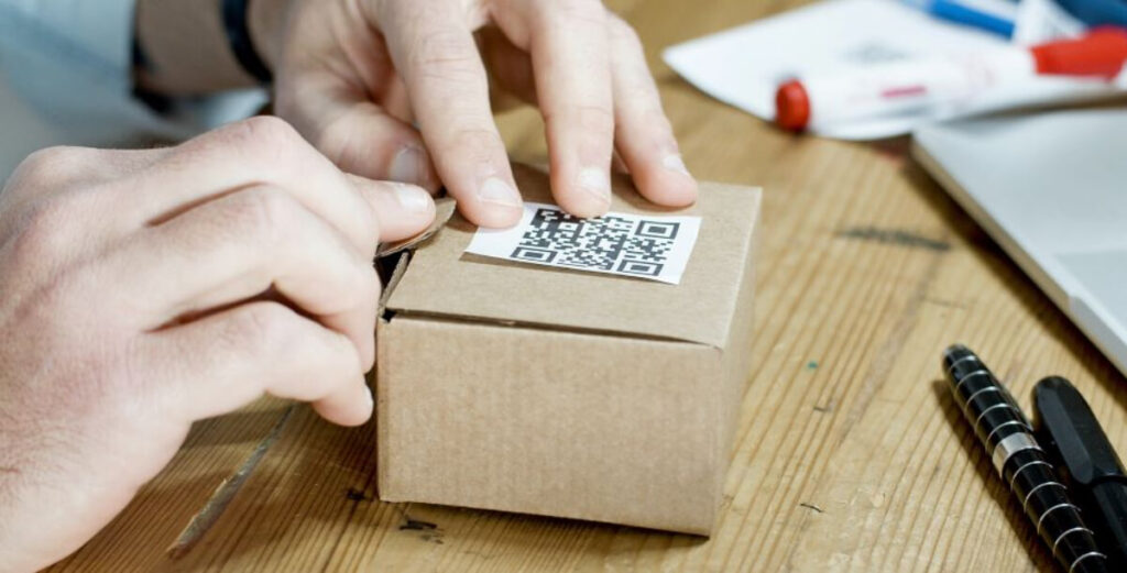 تصویر چسباندن برچسب QR روی جعبه مقوایی، مردی در حال چسباندن کد QR روی یک جعبه پستی ساده، نماد ترکیب طراحی سنتی و فناوری دیجیتال.